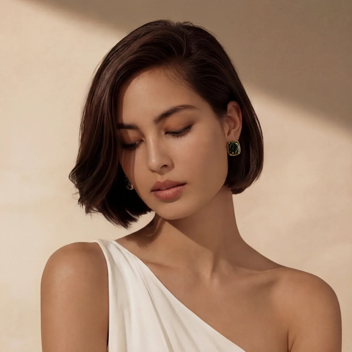 Woman wearing elegant green gemstone earrings with gold setting, styled with short bob and white one-shoulder top