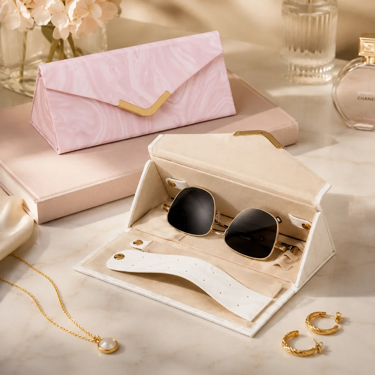 Elegant gold-framed sunglasses in a cream velvet case with pearl necklace and gold hoop earrings on marble