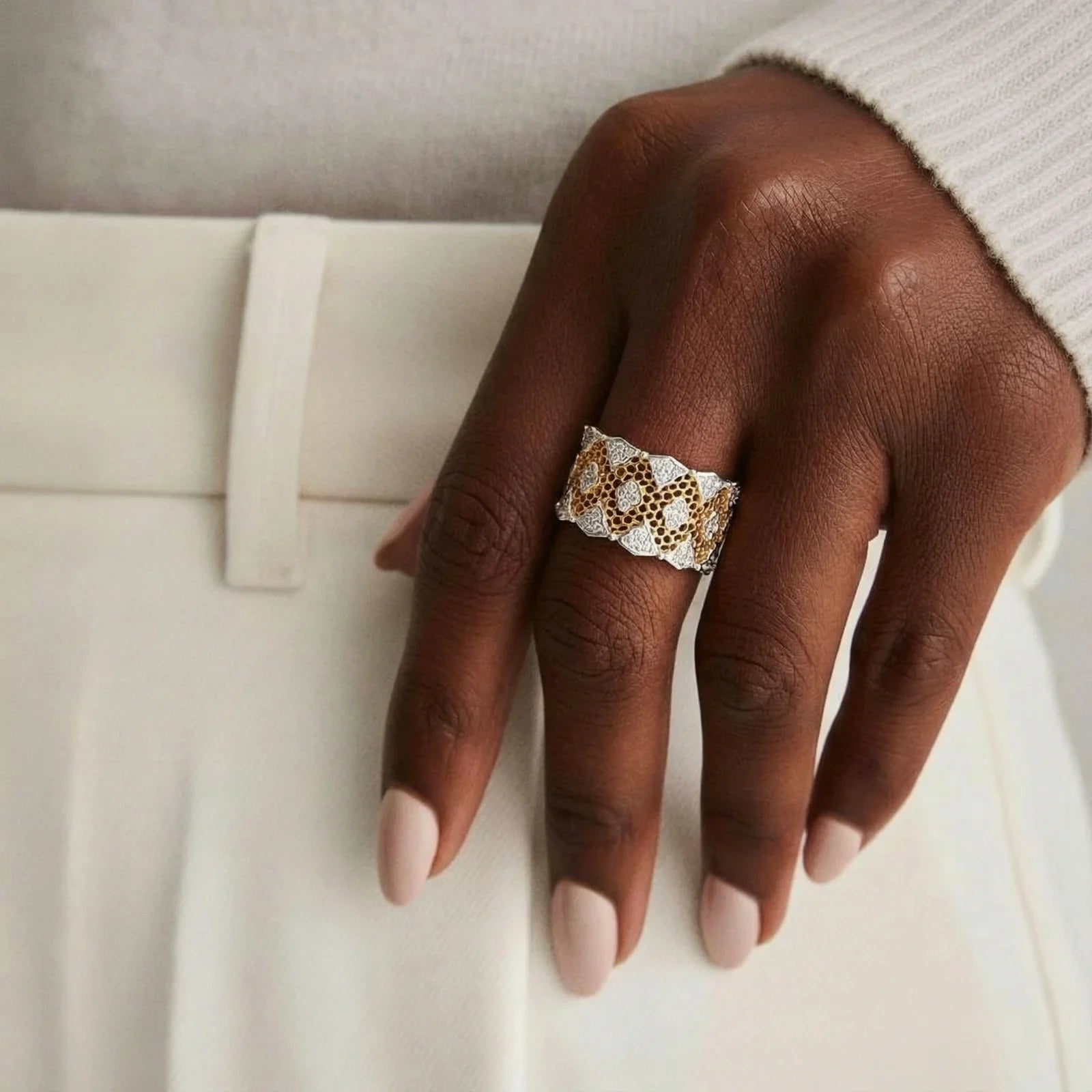 Elegant gold and silver statement ring with geometric pattern, worn on hand with neutral nails and white outfit.