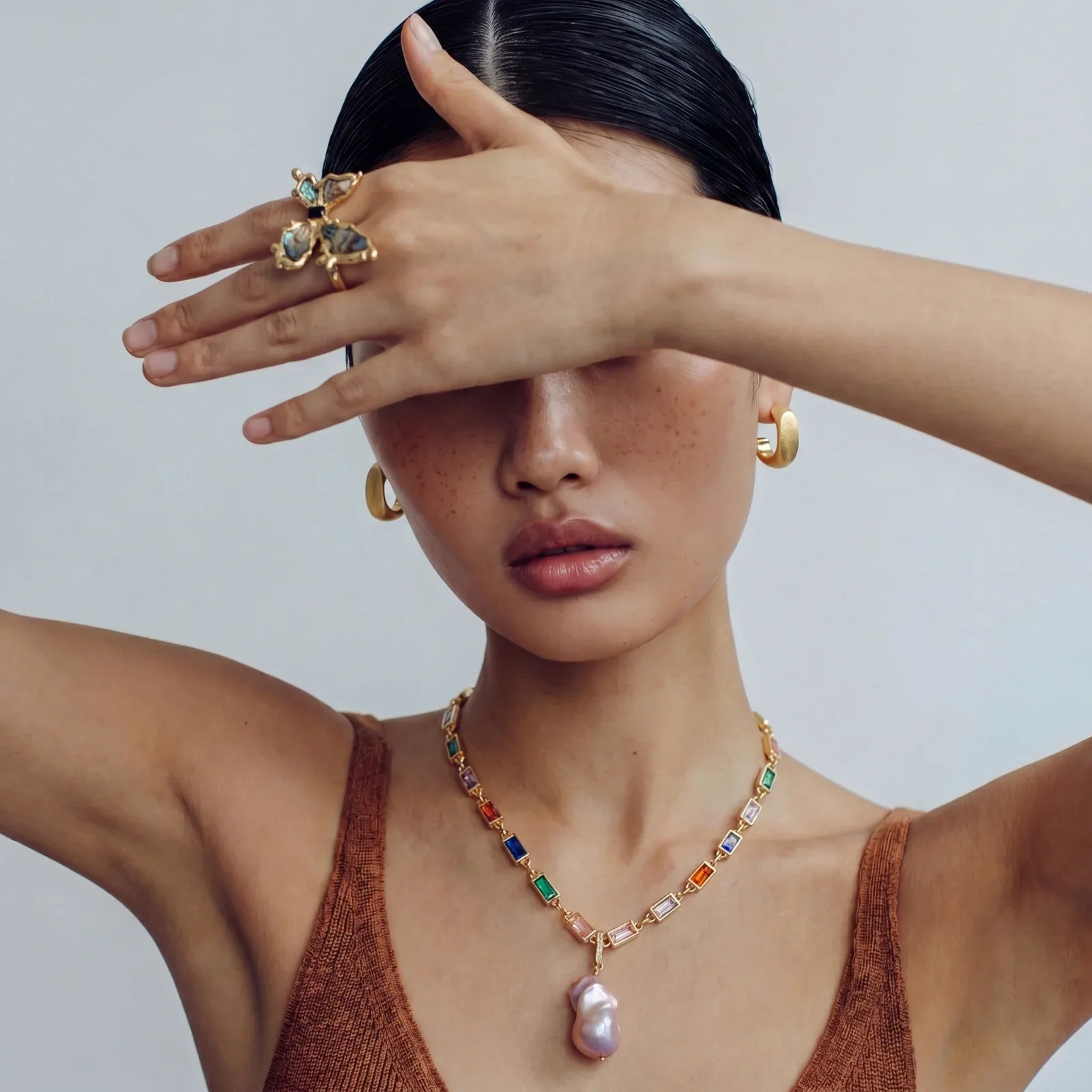 Woman wearing gold hoop earrings, colorful gemstone necklace with pearl pendant, and butterfly ring