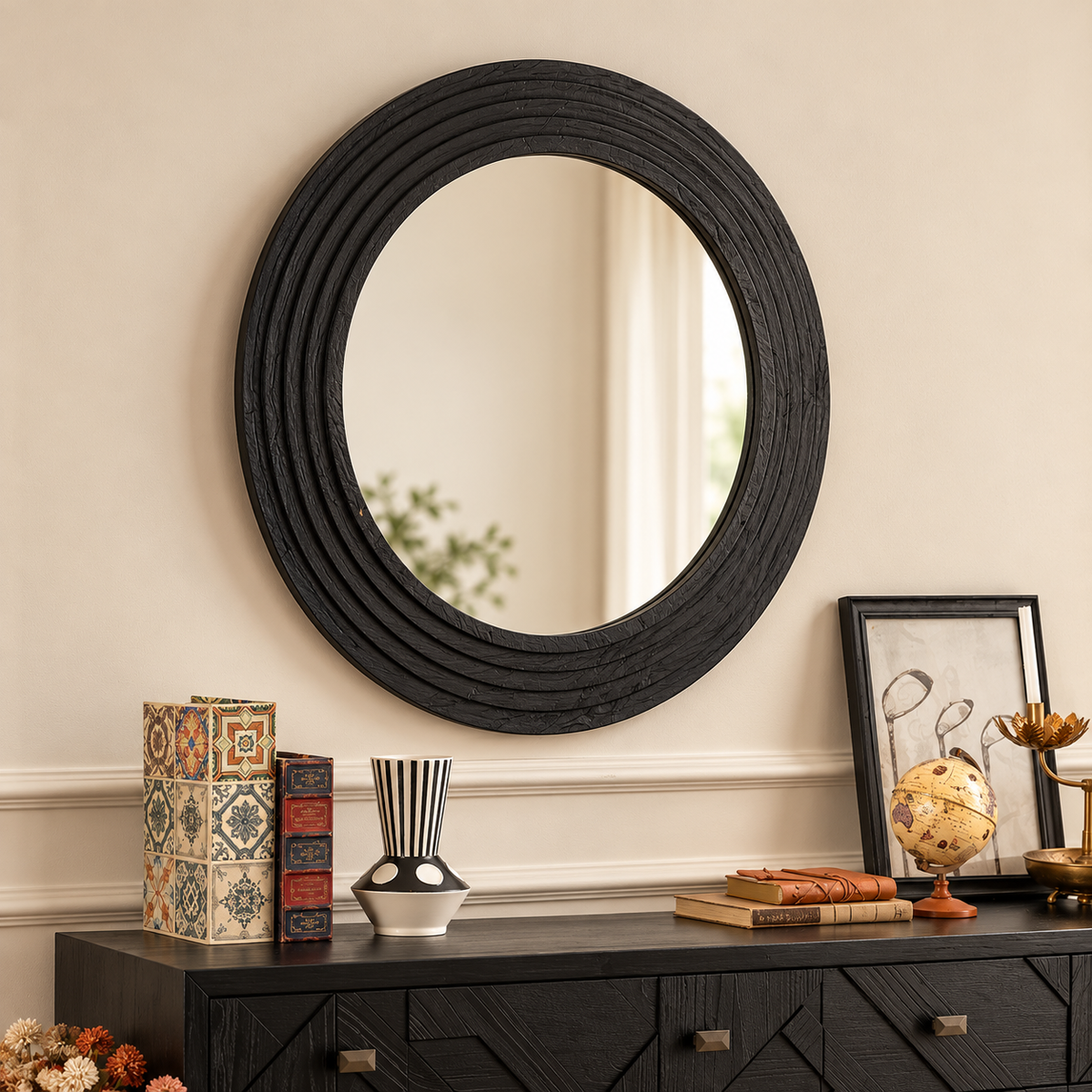 A large oval black-framed wall mirror with a textured, ribbed border hangs on a beige wall above a dark wood console table styled with books, a vase, and framed art.