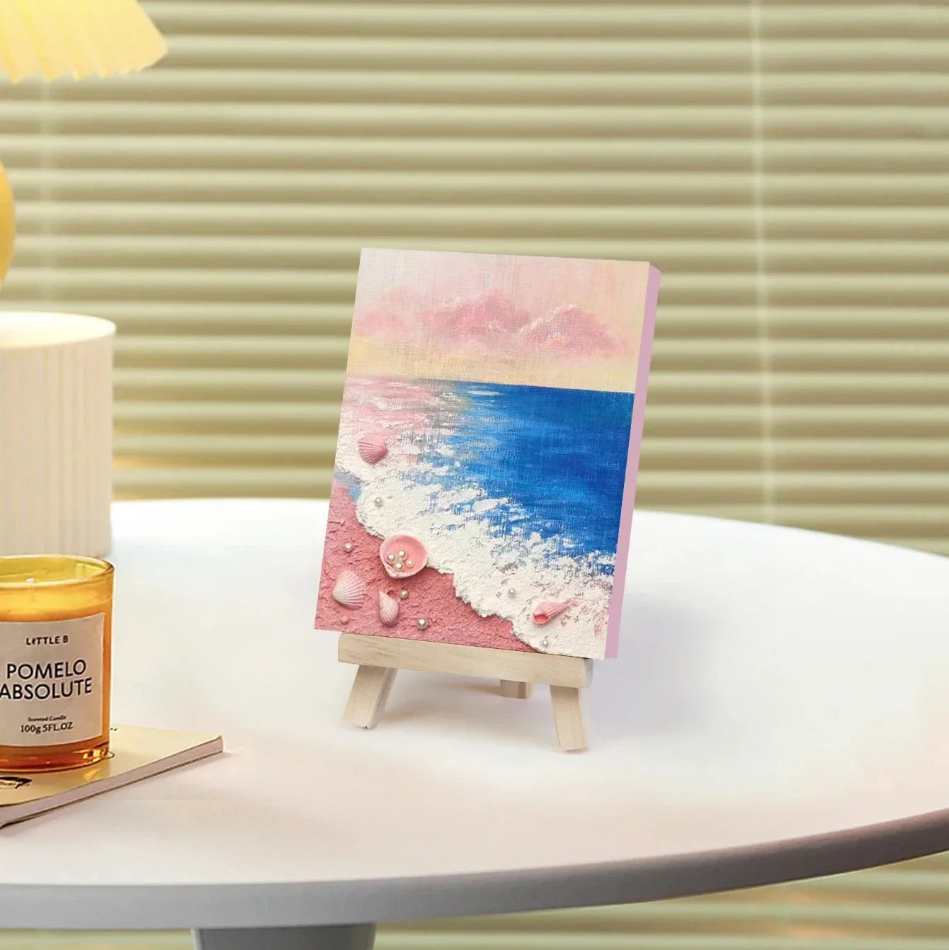 Mini easel displays pink ocean beach painting with seashells, on white table in modern decor